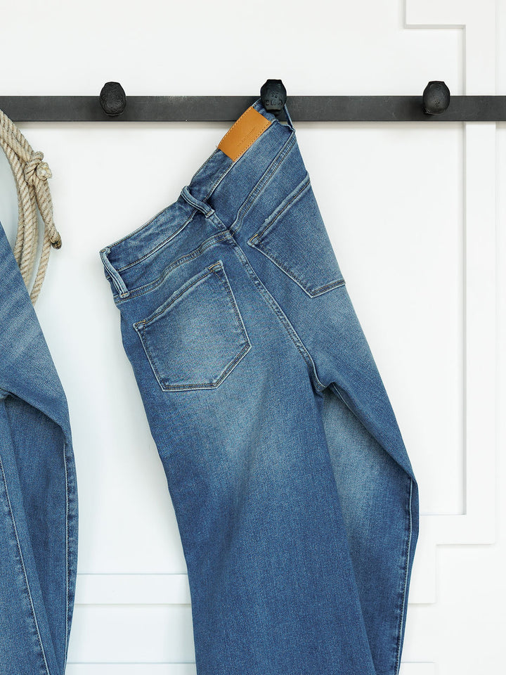 The Slayden High Rise Wide Leg Jean by Savannah Sevens western life{&}style, featuring a brown leather patch, hangs on a black wall hook against a white background; another pair of jeans is partially visible on the left.