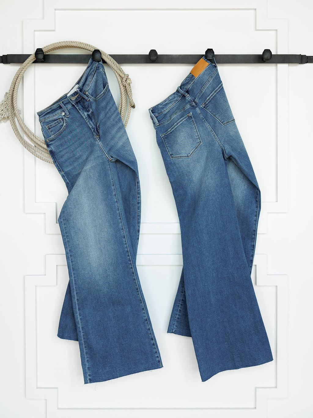 Two pairs of The Slayden High Rise Wide Leg Jean by Savannah Sevens western life{&}style hang side by side on a black rod against a white wall with decorative trim. A coiled beige rope hangs from the left end of the rod.