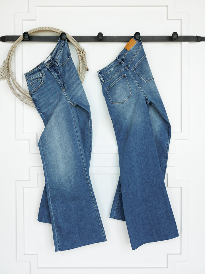 Two pairs of The Slayden High Rise Wide Leg Jean by Savannah Sevens western life{&}style hang side by side on a black rod against a white wall with decorative trim. A coiled beige rope hangs from the left end of the rod.