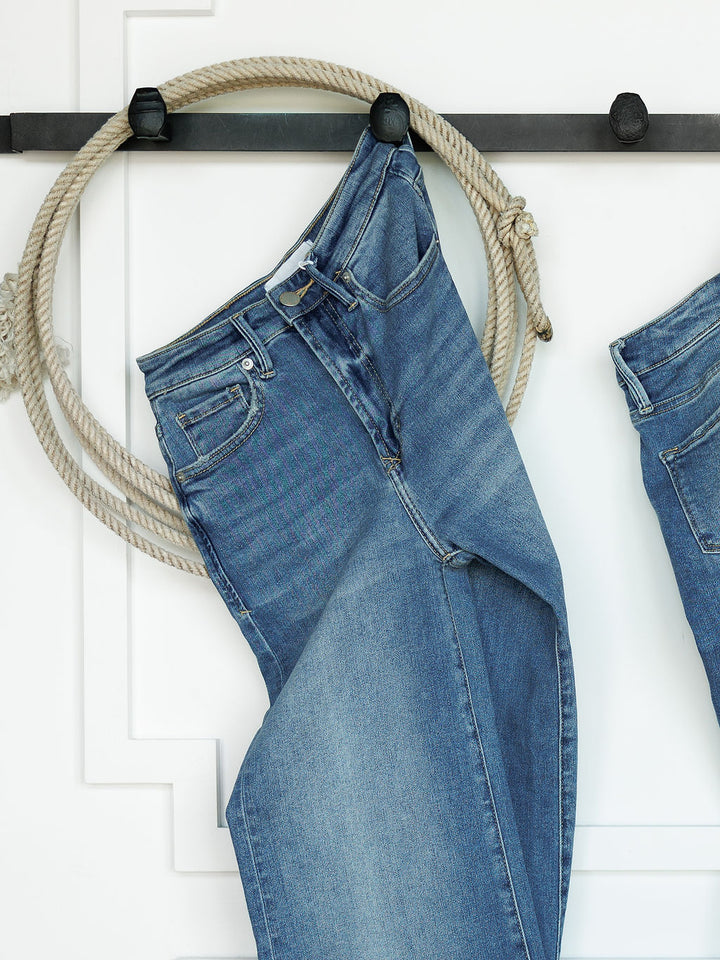 The Slayden High Rise Wide Leg Jean by Savannah Sevens western life{&}style hangs on a black wall hook draped over a coiled rope, set against a white wall with decorative trim.
