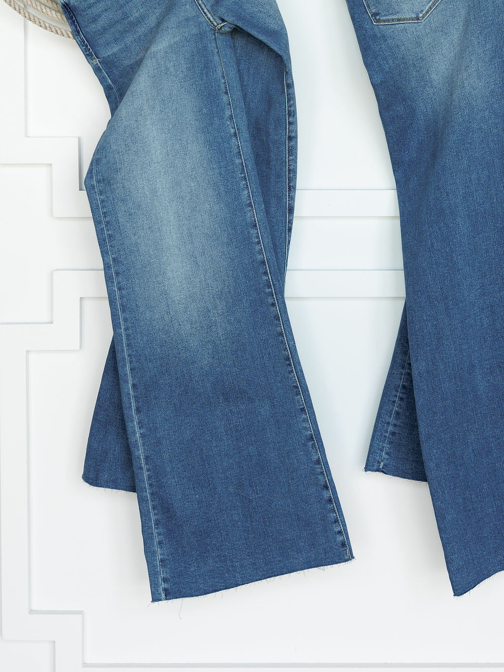 A close-up of The Slayden High Rise Wide Leg Jean by Savannah Sevens western life{&}style, featuring blue denim with a raw hem, hanging against a white textured wall.
