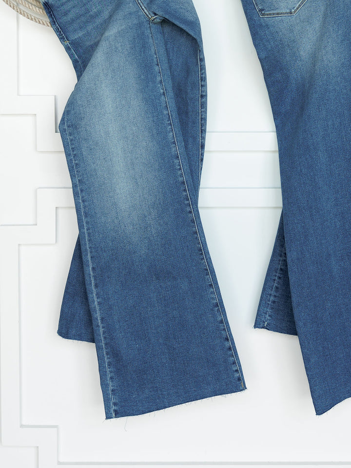 A close-up of The Slayden High Rise Wide Leg Jean by Savannah Sevens western life{&}style, featuring blue denim with a raw hem, hanging against a white textured wall.