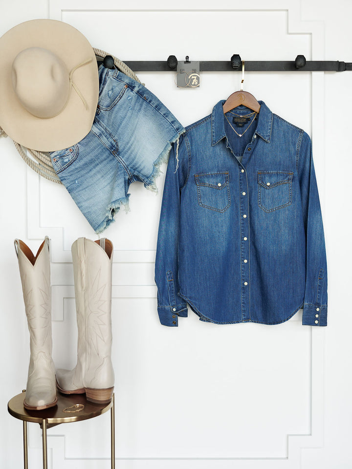 The Tekamah beige cowboy hat by Savannah Sevens western life{&}style, denim shorts, and a Pendleton denim shirt hang on a wall rack above tall white cowboy boots set on a small round table.
