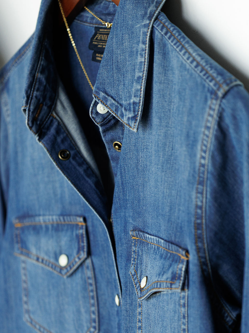 The Tekamah denim shirt by Savannah Sevens western life{&}style, featuring front snap pockets, hangs on a wooden hanger, partially unbuttoned.