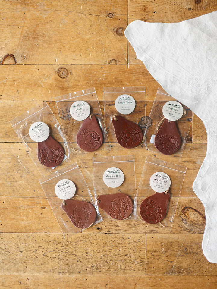 Seven Tooled Leather Car Fresheners by Savannah Sevens western life{&}style, each in teardrop shape with floral designs, are arranged on wood. Individually packaged in clear plastic with round labels. A white cloth is visible at top right.