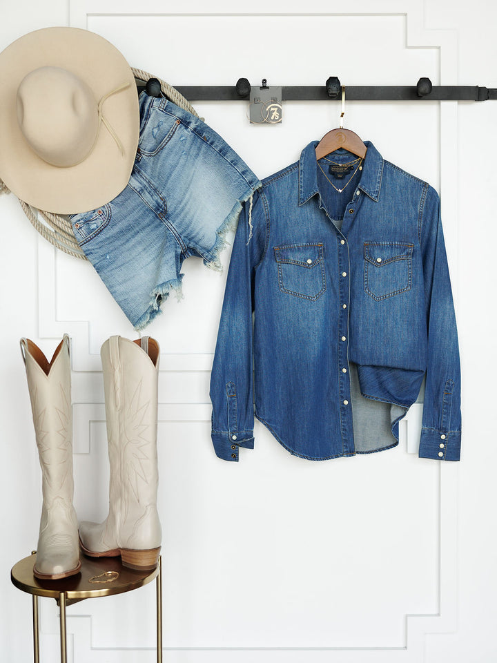 The Tekamah Savannah Sevens western life{&}style hangs on a wall rack besides frayed denim shorts a, with cream cowboy boots on a gold side table in front of a white paneled wall.