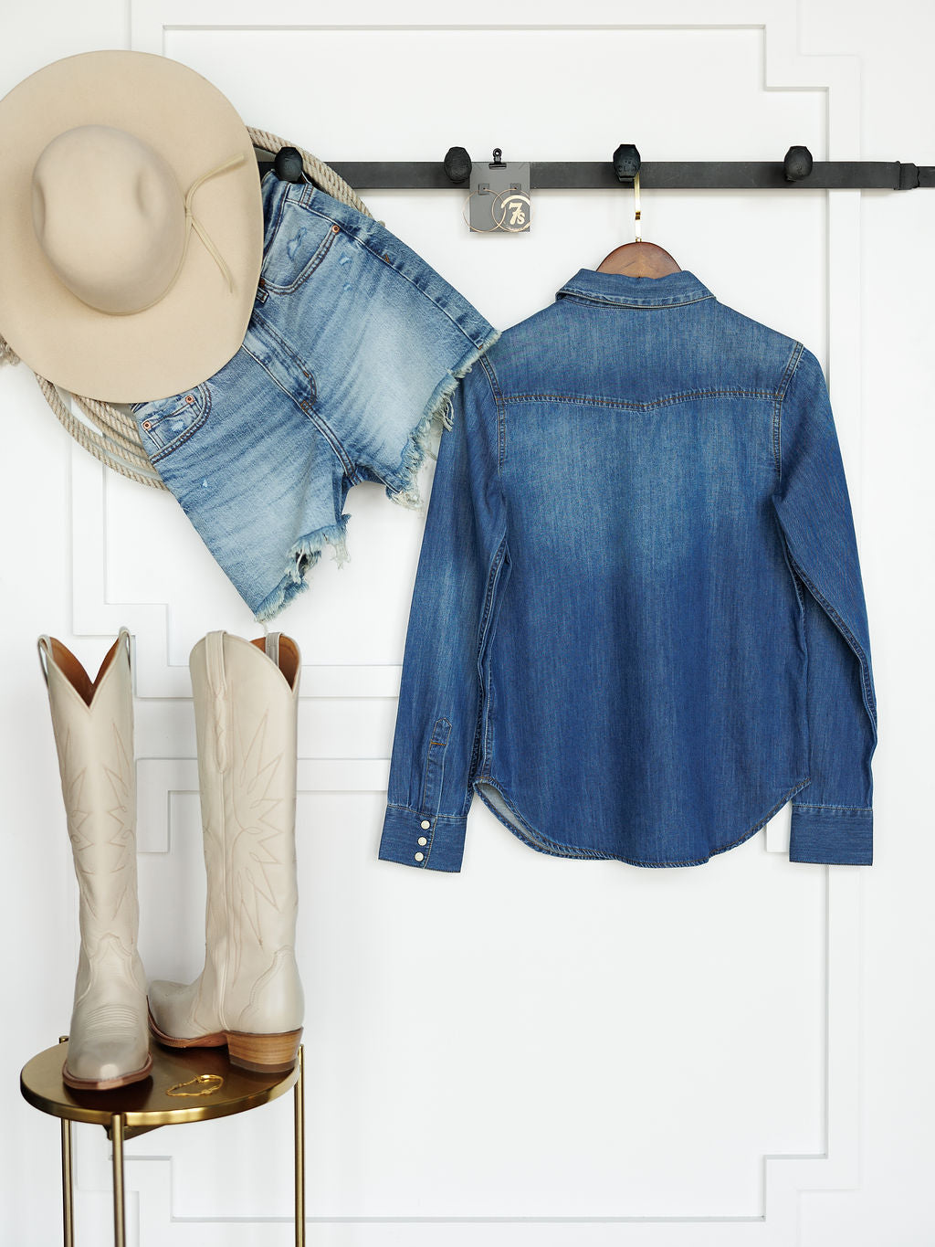 A pair of white cowboy boots by Savannah Sevens western life{&}style stand on a table, with denim shorts, a Pendleton denim shirt, and a beige cowboy hat hanging above against a white wall.