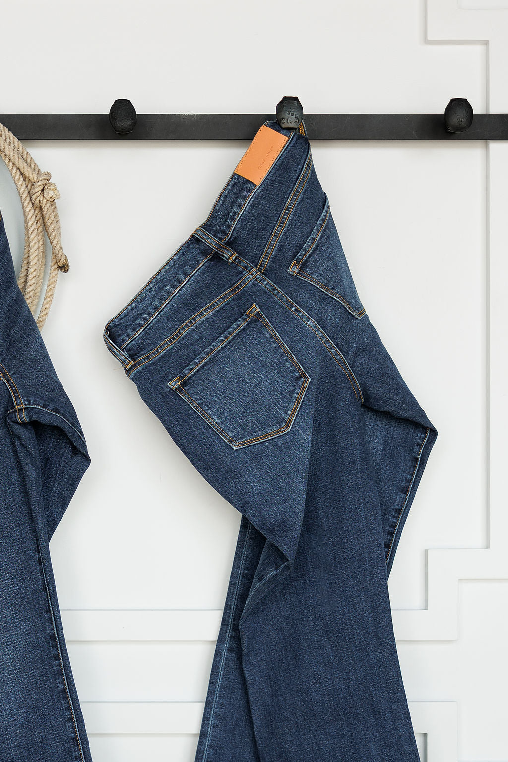 A pair of The Camryn Premier Rise Slim Straight Leg Jeans by Savannah Sevens western life{&}style hangs folded over a black wall hook, with a white paneled wall and part of a rope visible in the background.