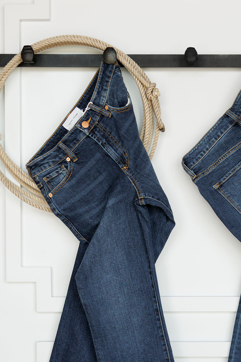 A pair of dark wash denim jeans hangs on a black rack with a coiled rope against a white wall. On the right, the Camryn Premier Rise Slim Straight Leg Jean by Savannah Sevens western life{&}style is partially visible.