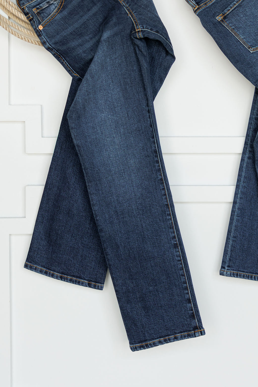 A pair of Savannah Sevens western life{&}style The Camryn Premier Rise Slim Straight Leg Jeans in dark wash denim hangs against a white paneled wall, displaying the jeans’ full length.