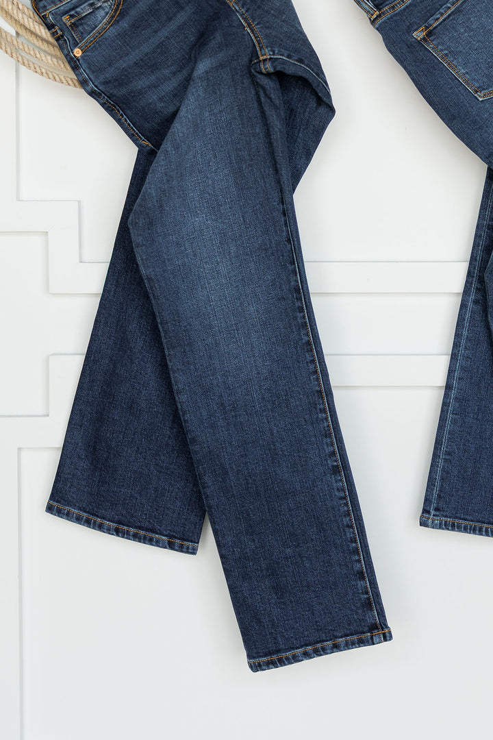 A pair of Savannah Sevens western life{&}style The Camryn Premier Rise Slim Straight Leg Jeans in dark wash denim hangs against a white paneled wall, displaying the jeans’ full length.