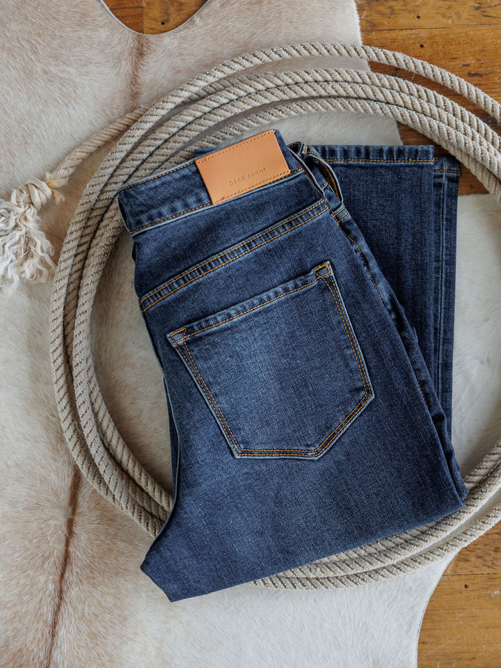 A pair of folded dark wash The Camryn Premier Rise Slim Straight Leg Jeans by Savannah Sevens western life{&}style, with a tan leather label, rests inside a coiled rope on a light animal hide rug atop a wooden floor.