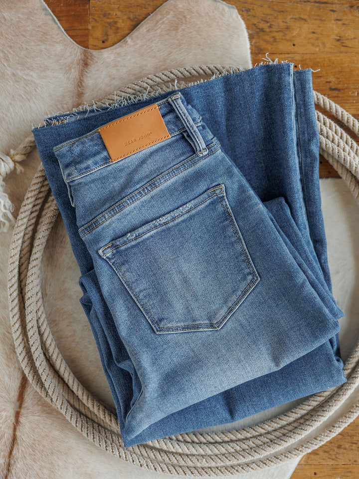 Two pairs of The Slayden High Rise Wide Leg Jeans by Savannah Sevens western life{&}style, one showing the back pocket with a brown leather patch, are neatly folded on a cowhide rug with a coiled rope around them on a wooden floor.