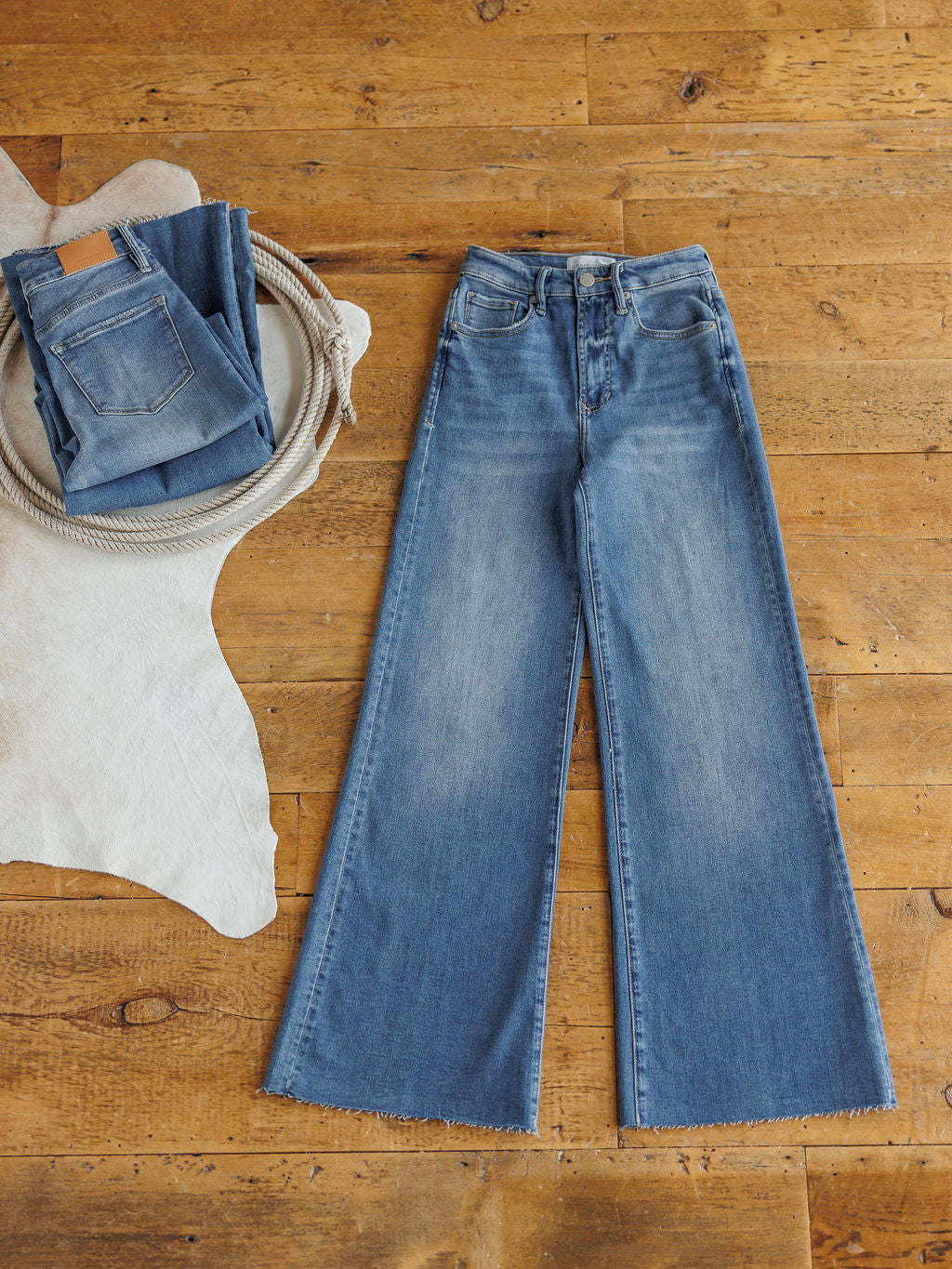 The Slayden High Rise Wide Leg Jean by Savannah Sevens western life{&}style is laid flat on a wooden floor. A folded pair rests on a round tray with coiled rope, beside a white cowhide rug.