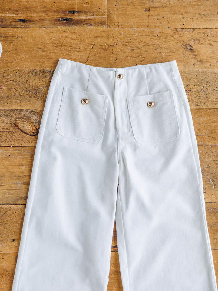 The Owasso white denim wide leg crop pants by Savannah Sevens western life{&}style, featuring two front pockets and gold buttons, are laid flat on a wooden floor.