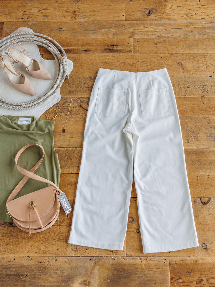 Flat lay of The Owasso white denim wide leg crop pants by Savannah Sevens western life{&}style on a wooden floor, styled with a green top, tan crossbody bag, beige pointed-toe shoes, and a chic coiled rope accessory.