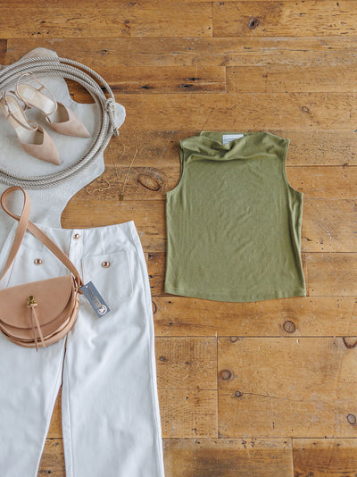 Flat lay of The Mandan olive cowl neck sleeveless top by Savannah Sevens western life{&}style, paired with white pants with gold buttons, a beige crossbody bag, beige pointed flats, and gold hoop earrings on a wooden floor.