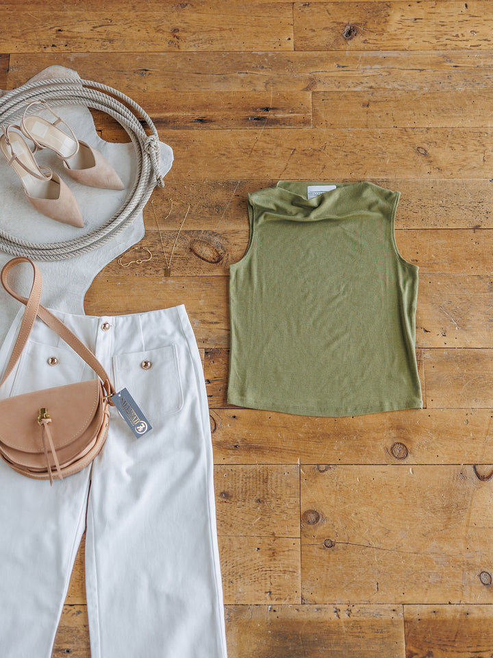 Flat lay of The Mandan olive cowl neck sleeveless top by Savannah Sevens western life{&}style, paired with white pants with gold buttons, a beige crossbody bag, beige pointed flats, and gold hoop earrings on a wooden floor.
