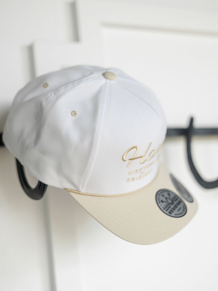 A Hooey Benton Cap by Savannah Sevens western life{&}style hangs on a black hook. This white and beige trucker cap features gold embroidery, an adjustable snapback, and a round black sticker on the beige brim against a white paneled wall.
