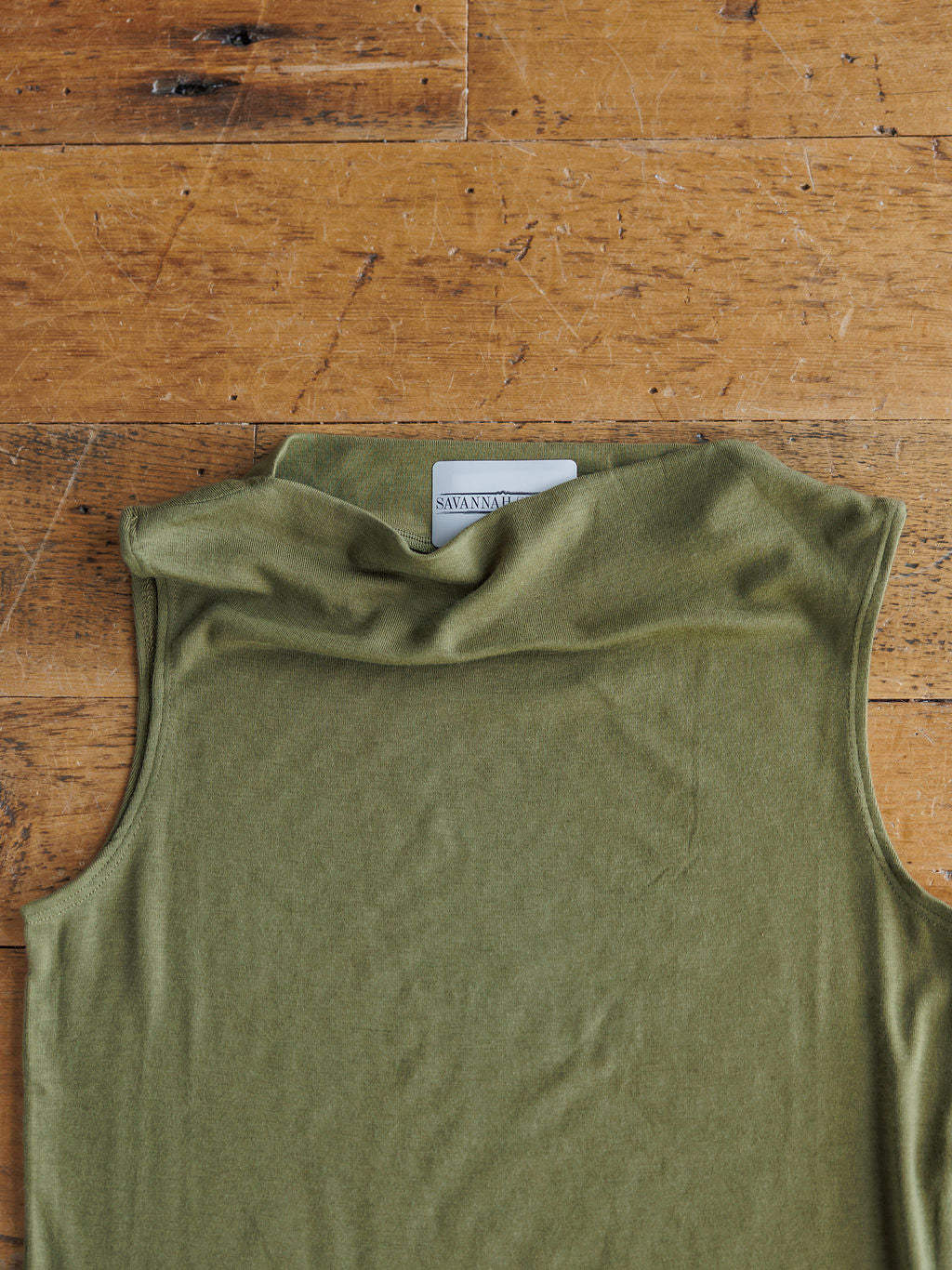 The Mandan by Savannah Sevens western life{&}style, a sleeveless olive cowl neck tri-blend top, is laid flat on a wooden surface with its inner label partially visible.