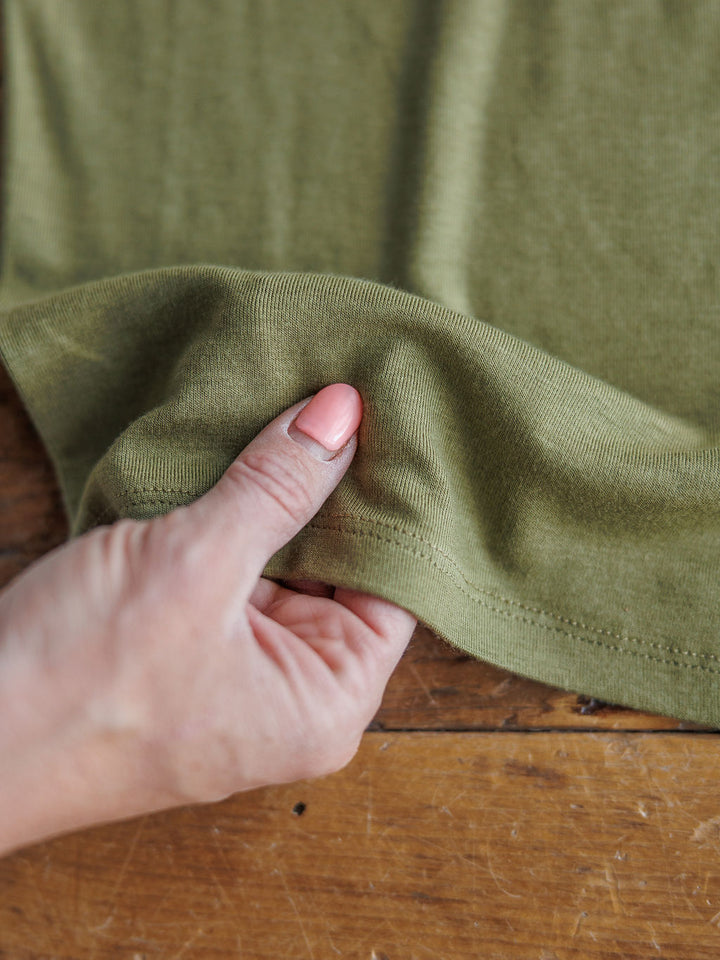 A hand with neatly manicured nails gently pinches the edge of The Mandan olive cowl neck top by Savannah Sevens western life{&}style, made from tri-blend fabric and resting on a wooden surface.