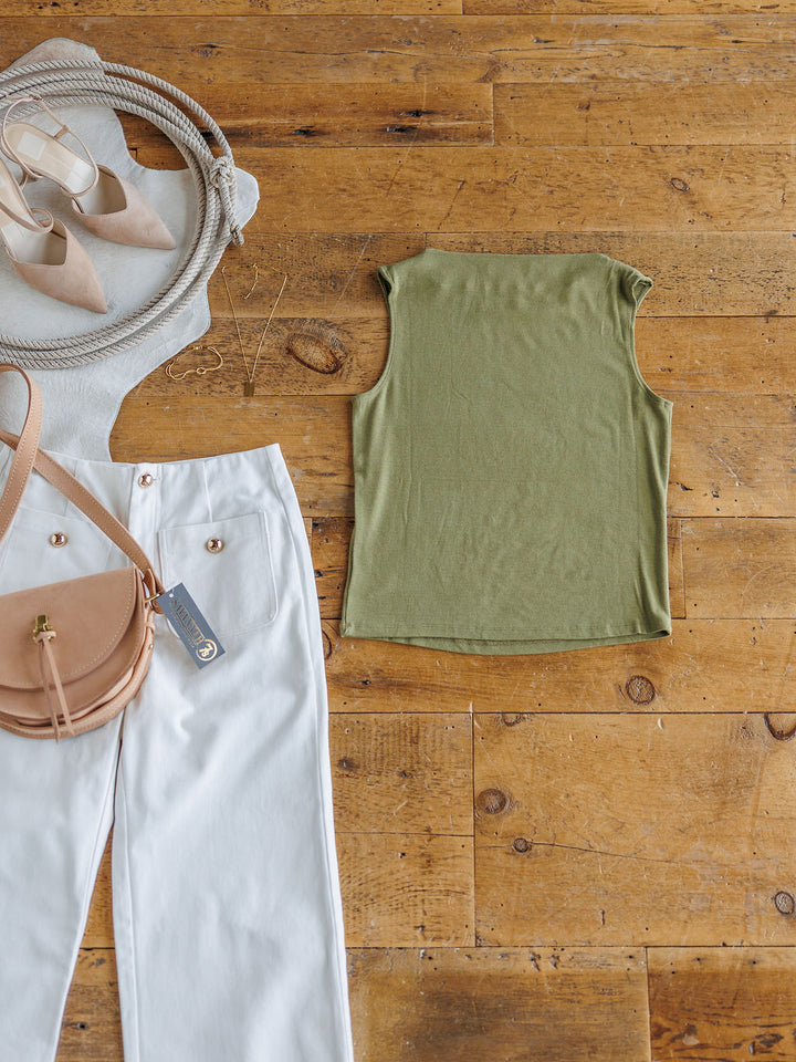 Flat lay of The Mandan olive cowl neck top by Savannah Sevens western life{&}style, white pants with gold buttons, a tan crossbody bag, beige pointed-toe heels, and a white round straw bag on a wooden floor.