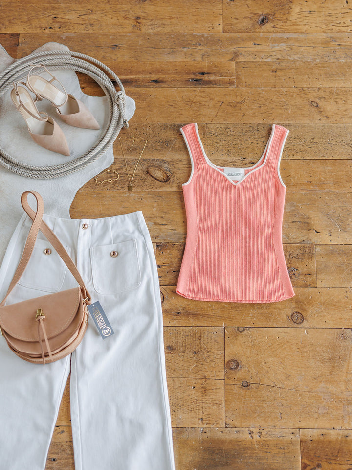Flat lay of The Lasara by Savannah Sevens western life{&}style: coral ribbed sleeveless top, white high-waisted pants, tan crossbody bag, beige pointed-toe heels, gold jewelry, and a woven gray hat on a wooden floor.