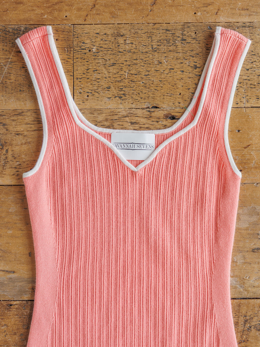 The Lasara by Savannah Sevens western life{&}style is a sleeveless pink ribbed knit dress with white trim on the v-neck and armholes, shown on a wooden surface.
