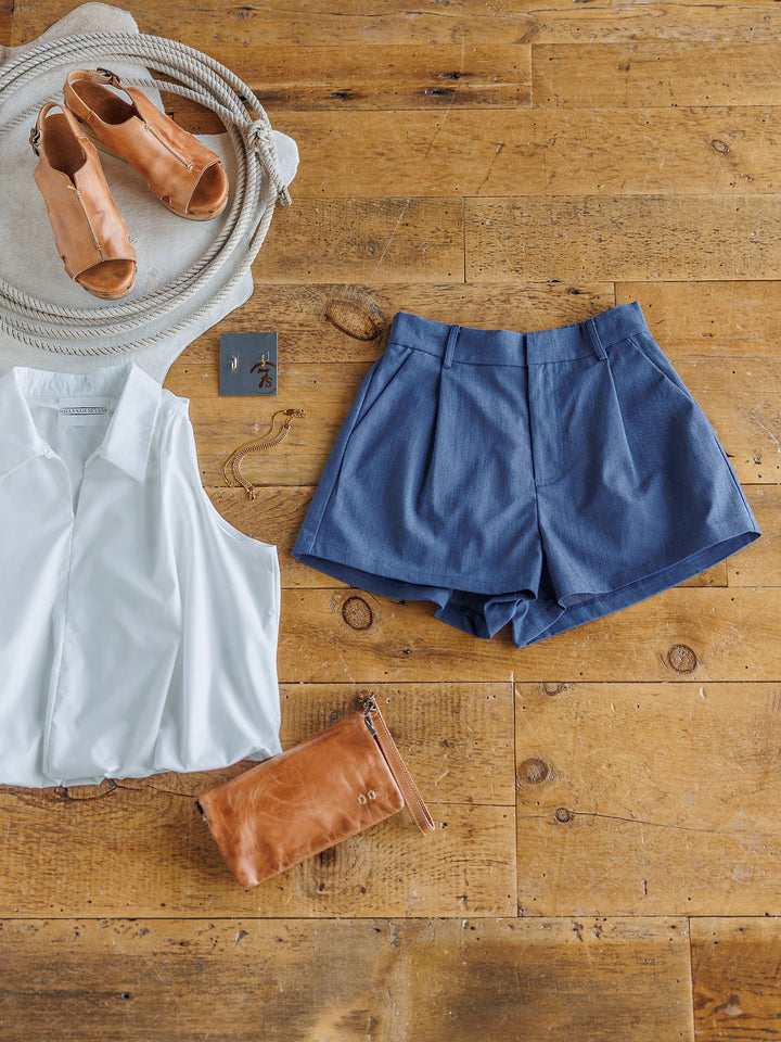 A flat lay on a wooden floor featuring The Lenox white sleeveless blouse by Savannah Sevens western life{&}style, high waisted denim blue shorts, tan heeled sandals, a tan clutch, a coiled rope, and a small square card.