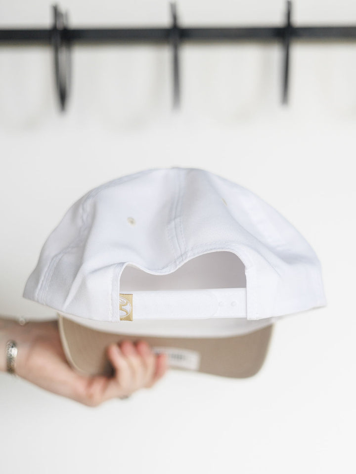 A hand holds a white Hooey Benton Cap by Savannah Sevens western life{&}style upside down, showcasing its adjustable snap back strap. The blurred background includes a black rack.
