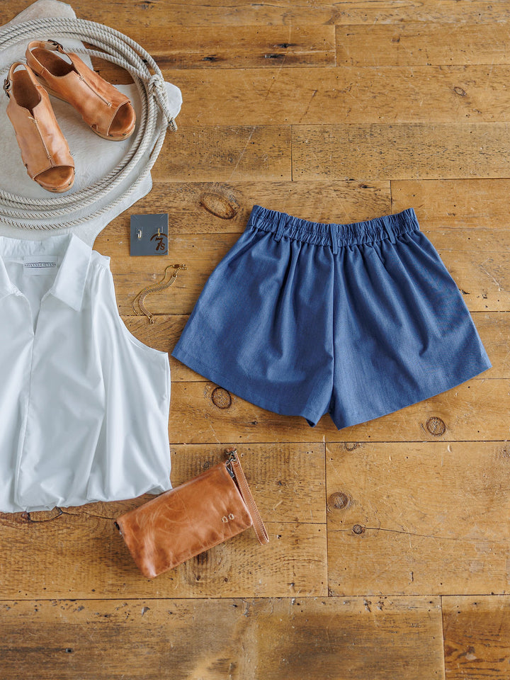 Flat lay of The Lenox by Savannah Sevens western life{&}style: sleeveless white blouse, cotton blend denim blue elastic waist shorts, tan sandals, tan clutch, necklace, and square keychain on a wooden floor.