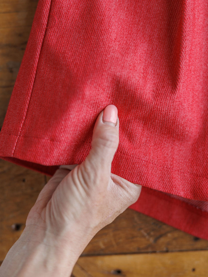 A hand holds the hem of The Ralston, red high-waisted cotton blend shorts by Savannah Sevens western life{&}style, displayed on a wooden surface.