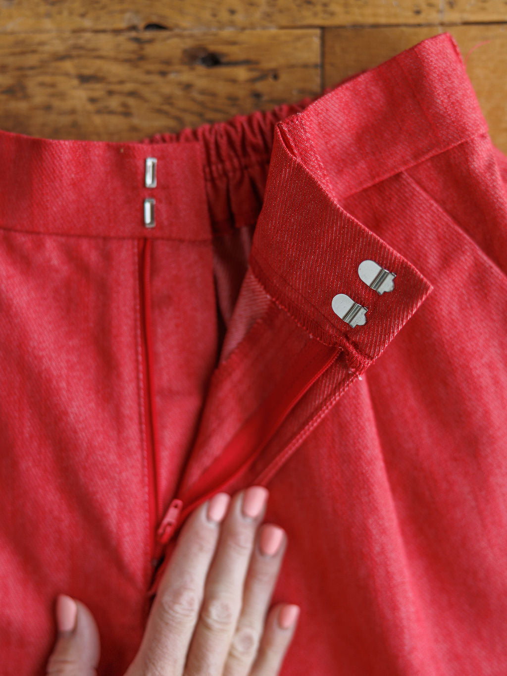 A close-up of a hand with light pink nails holding the waistband of The Ralston, high waisted tomato red shorts by Savannah Sevens western life{&}style, featuring a zipper and two metal hook-and-bar fasteners on a wooden surface.