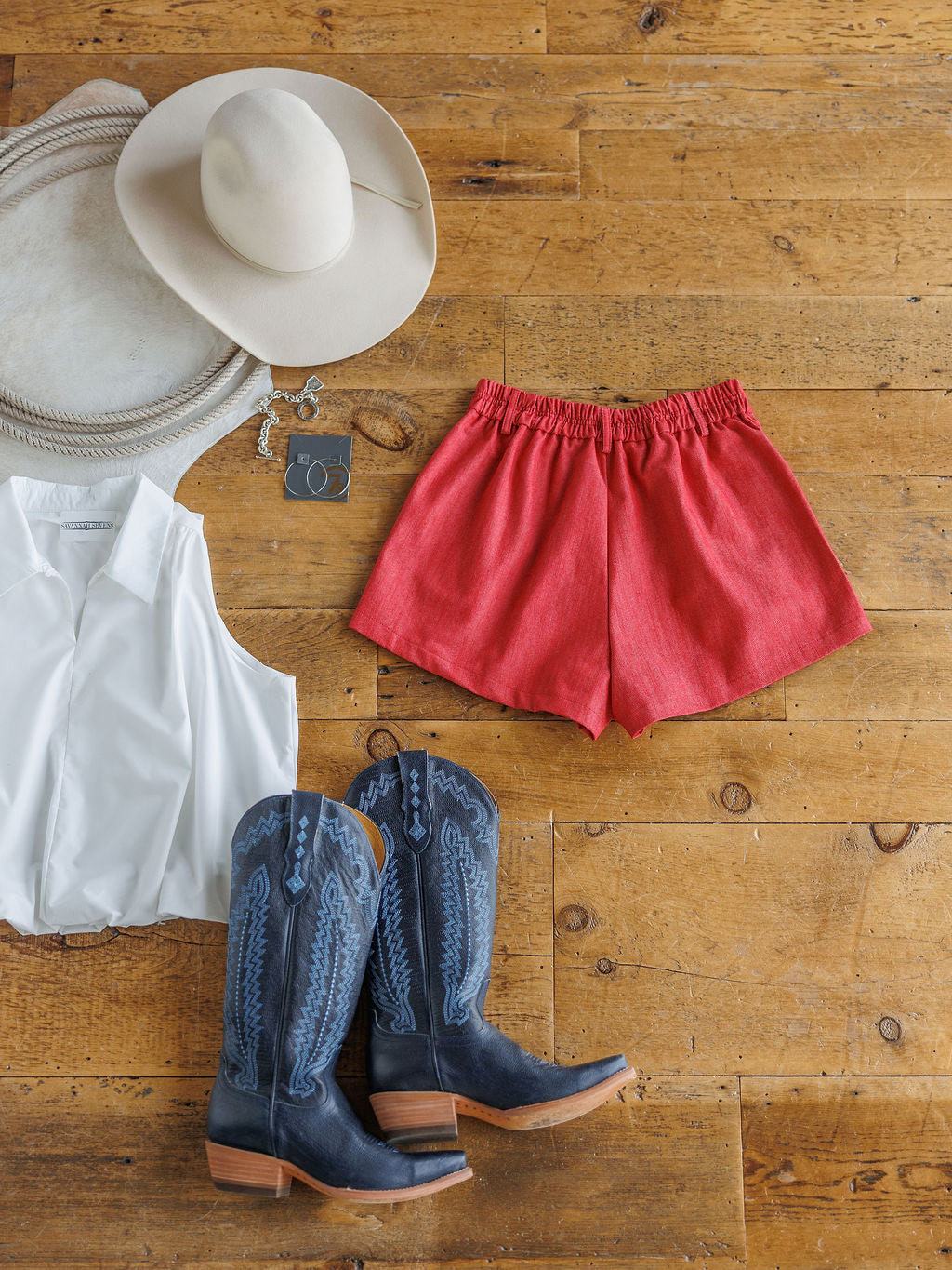 A flat lay of The Ralston by Savannah Sevens western life{&}style—a white sleeveless blouse with red high-waisted cotton blend shorts, blue cowboy boots, beige cowboy hat, coiled rope, and keys on a wooden floor.
