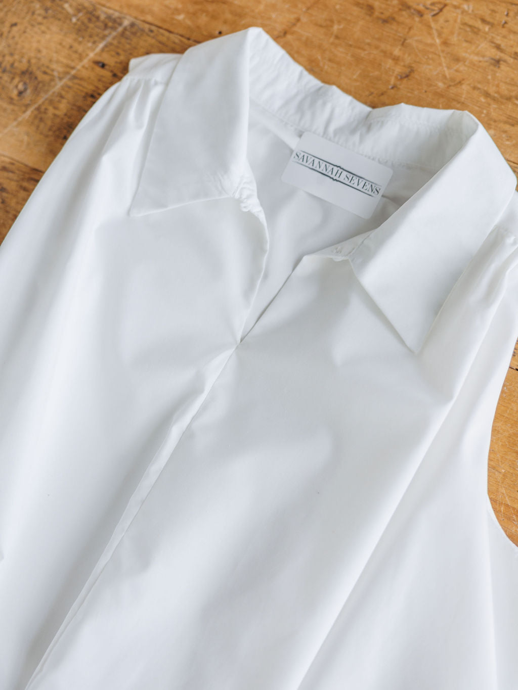 A sleeveless white top with a crisp texture and collared neckline, The Panora by Savannah Sevens western life{&}style, is laid neatly on a wooden surface, showcasing its clean cotton blend appeal.