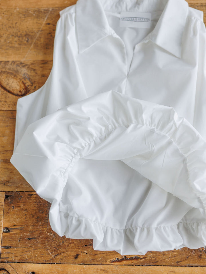 The Panora by Savannah Sevens western life{&}style, a sleeveless white top with a ruffled elastic bubble hem, is displayed on a rustic wooden surface.