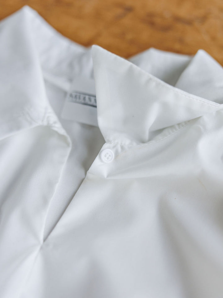A close-up of The Panora, a crisp sleeveless white cotton-blend top by Savannah Sevens western life{&}style, featuring a button near the neckline and slightly open collar, neatly pressed on a light brown textured surface.