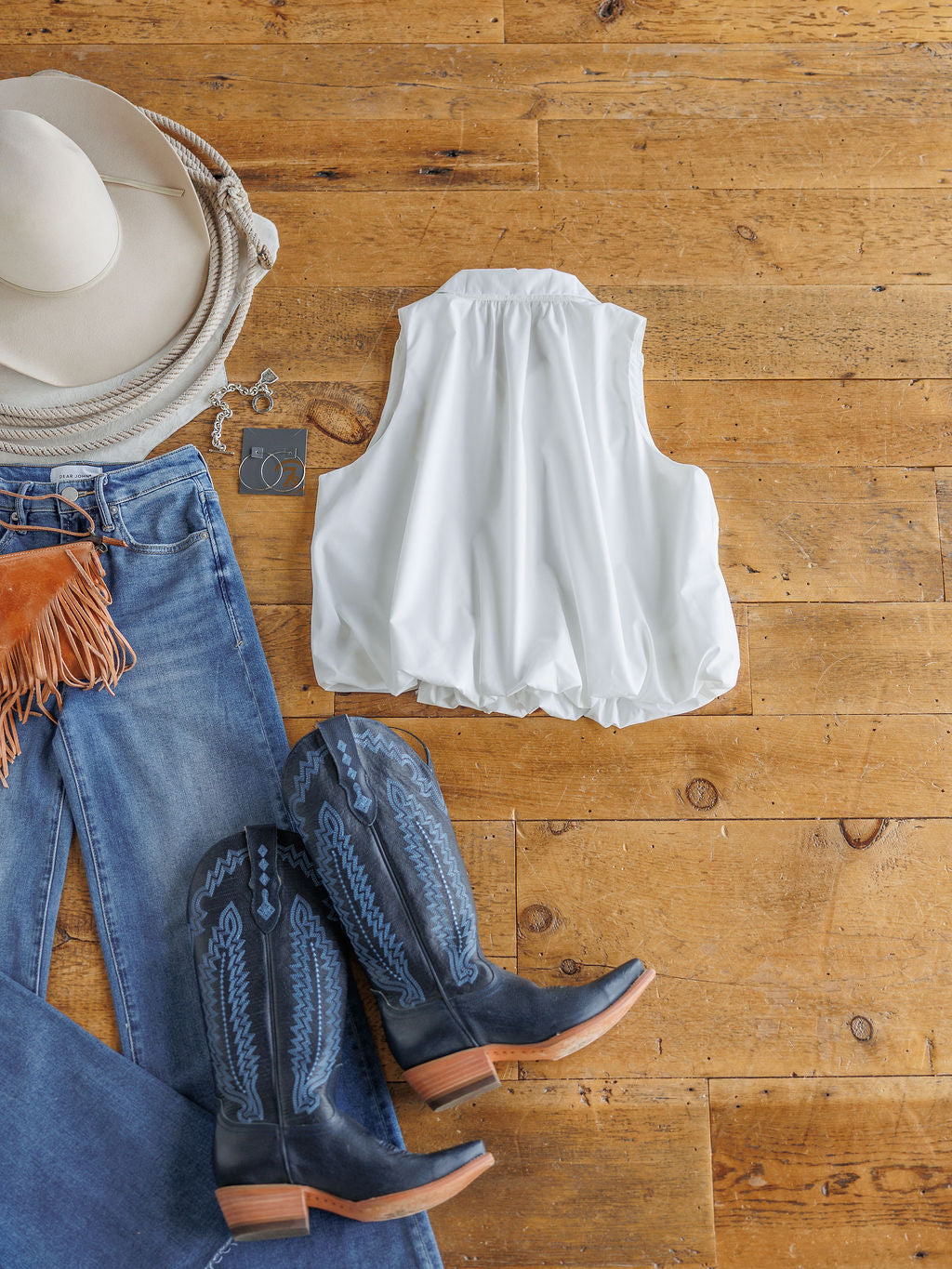 Flat lay of The Panora by Savannah Sevens western life{&}style: a sleeveless white top styled with blue fringe jeans, black and blue cowboy boots, a white cowboy hat, and silver hoop earrings on a wooden floor.