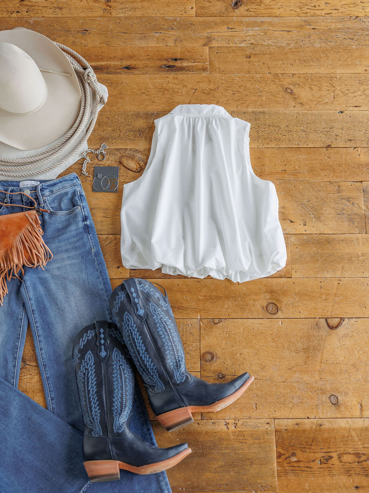 Flat lay of The Panora by Savannah Sevens western life{&}style: a sleeveless white top styled with blue fringe jeans, black and blue cowboy boots, a white cowboy hat, and silver hoop earrings on a wooden floor.