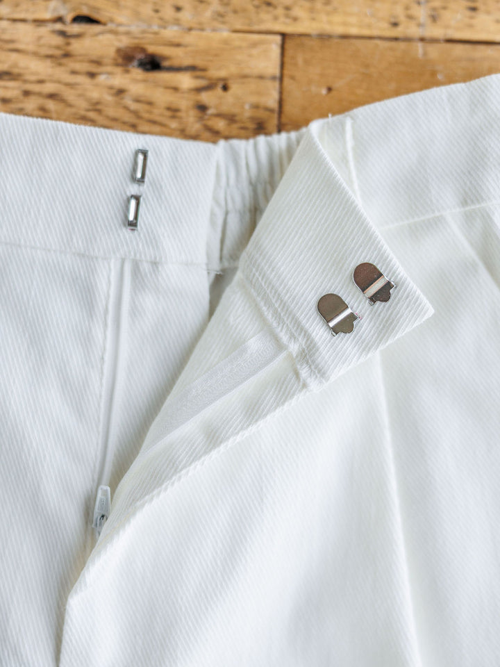 Close-up of the waistband of The Morley by Savannah Sevens western life{&}style—white pleated shorts with a zipper, dual hook-and-bar fasteners, and subtle pinstripe texture, laid on a wooden surface.