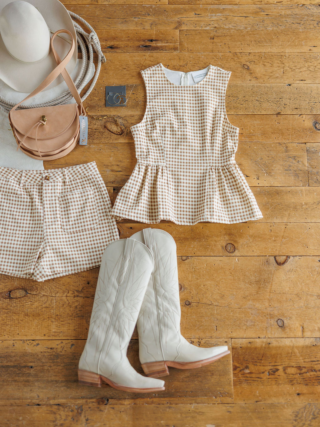 Flat lay of The Minetto Set by Savannah Sevens western life{&}style—beige gingham sleeveless top and high-waisted shorts, styled with white cowboy boots, round beige crossbody bag, wide-brimmed hat, and silver bracelet on a wooden floor.