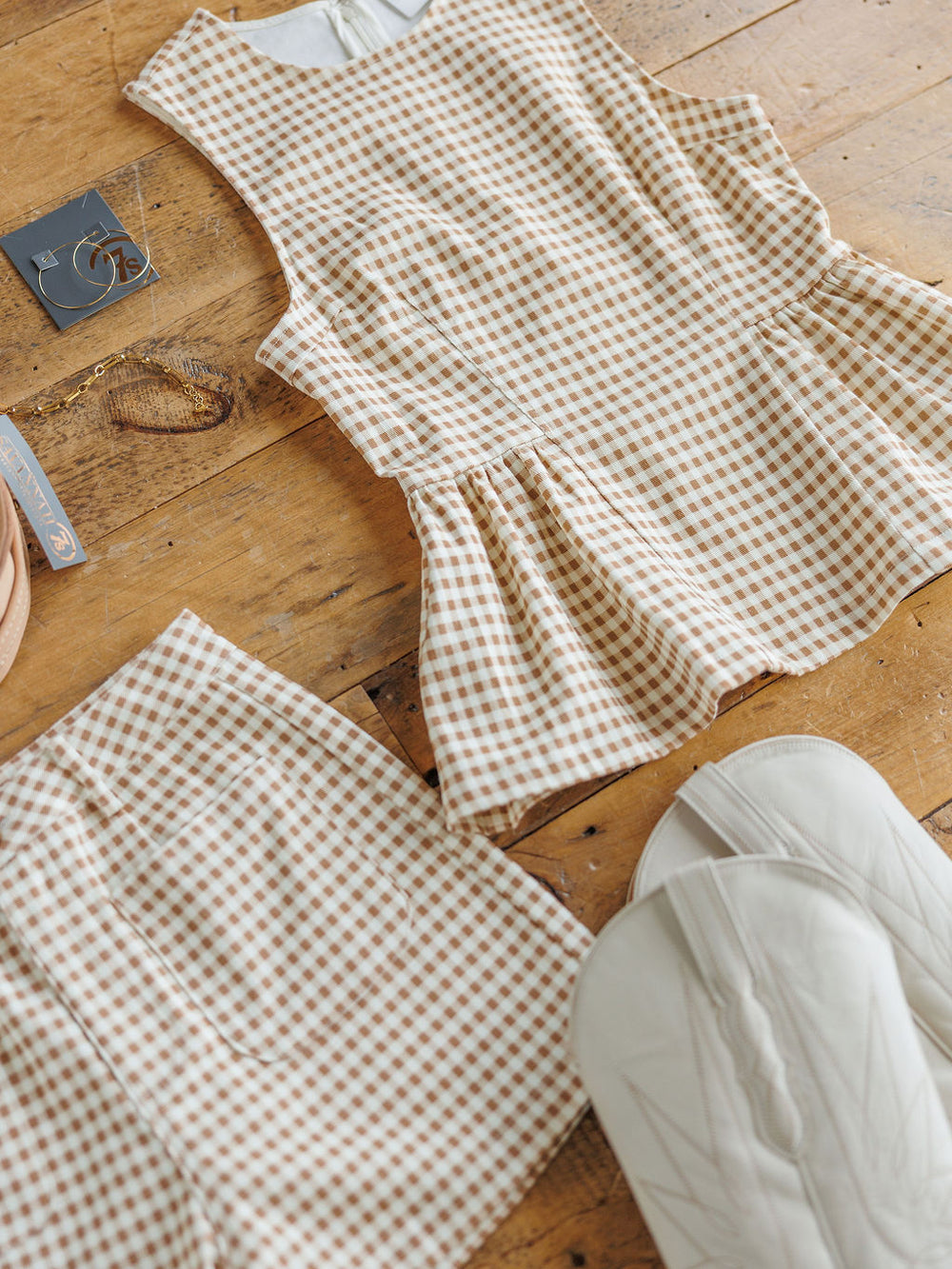The Minetto {Set} by Savannah Sevens western life{&}style features a sleeveless beige and white checkered peplum top with matching high-waisted shorts, styled with white quilted accessories, gold jewelry, and earrings on a wooden floor.