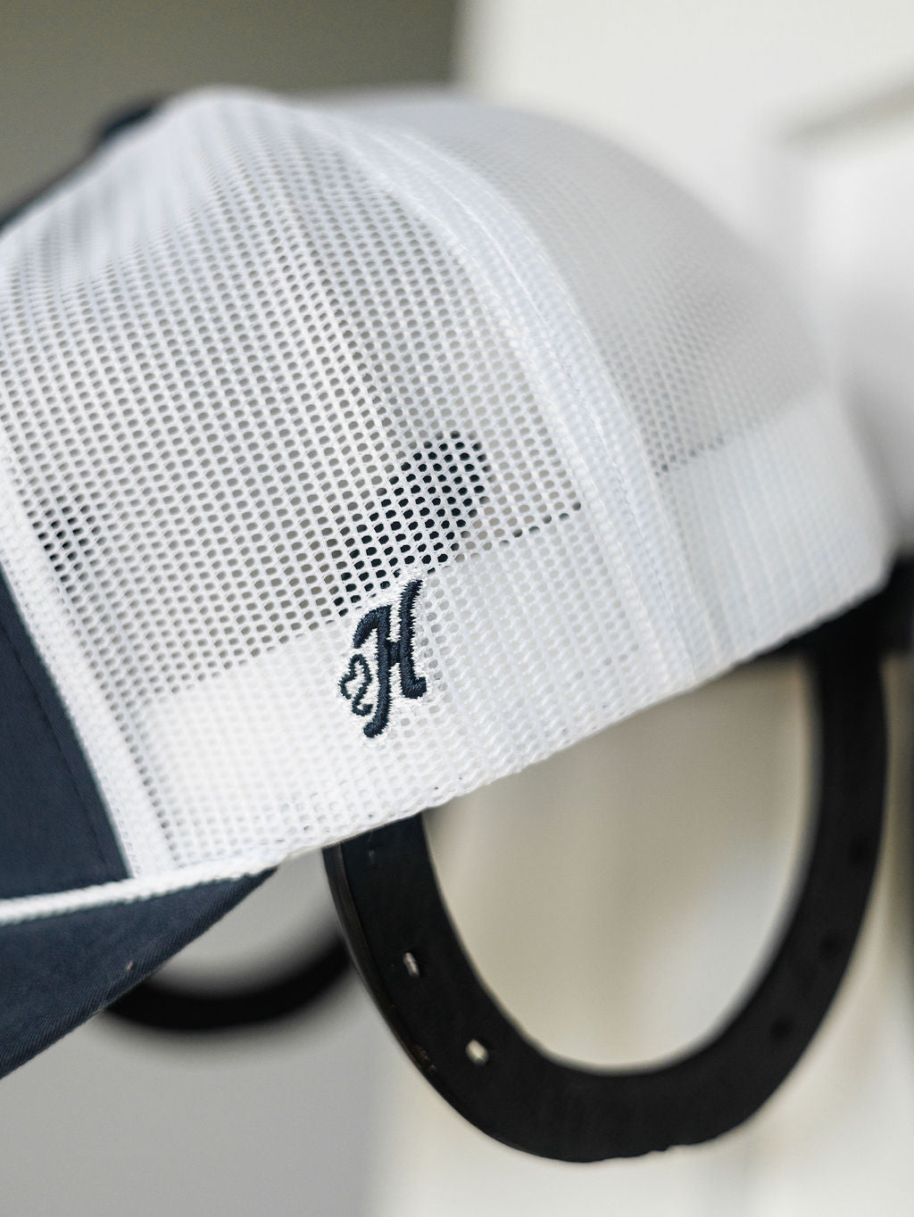 The Hooey Colby Cap by Savannah Sevens western life{&}style is a white mesh trucker cap with a black embroidered H, shown on a black horseshoe-shaped hook. It features an adjustable snap back for comfort.