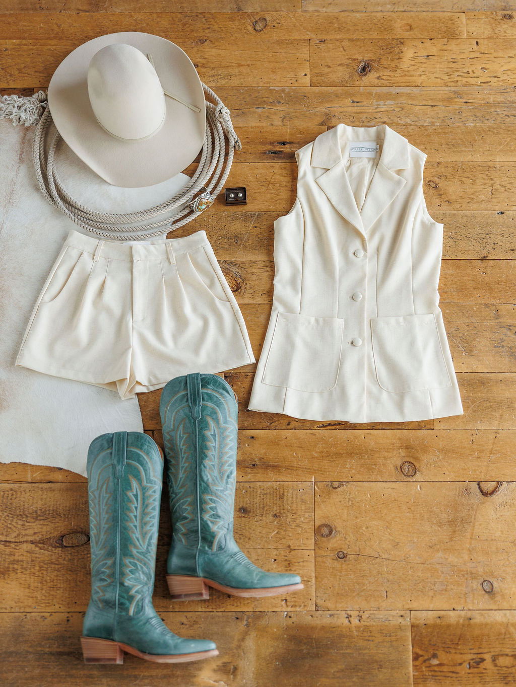Flat lay of The Savona {Set} by Savannah Sevens western life{&}style—featuring a cream cowboy hat, sleeveless blazer, linen-blend shorts, and teal cowboy boots on a rustic wooden floor.