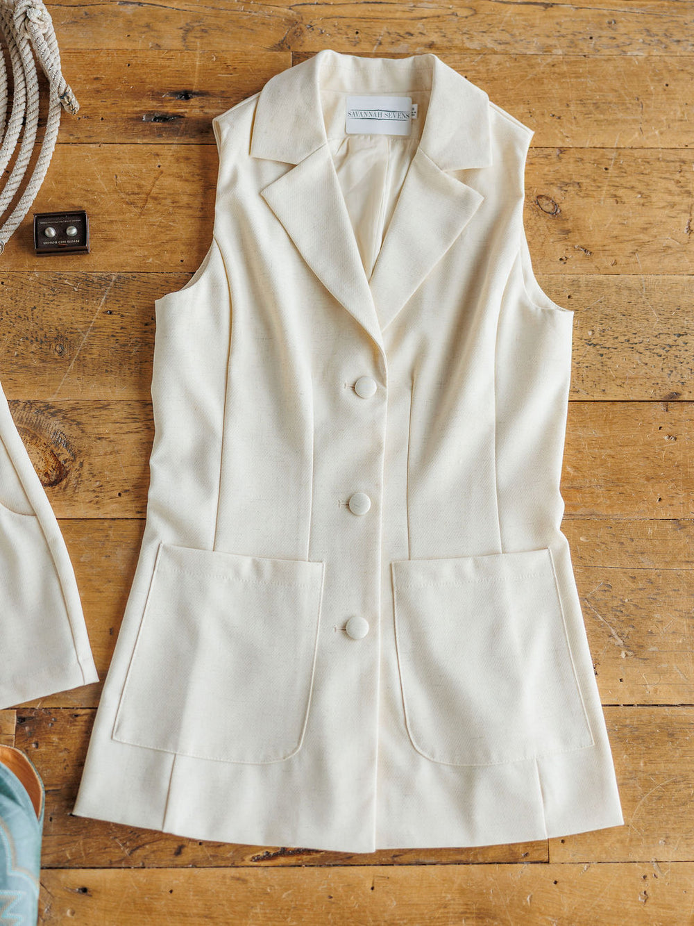 The Savona Set by Savannah Sevens western life{&}style: a sleeveless, cream vest with collar, 3 front buttons, and two large pockets—ideal with linen blend shorts—displayed flat on rustic wood.