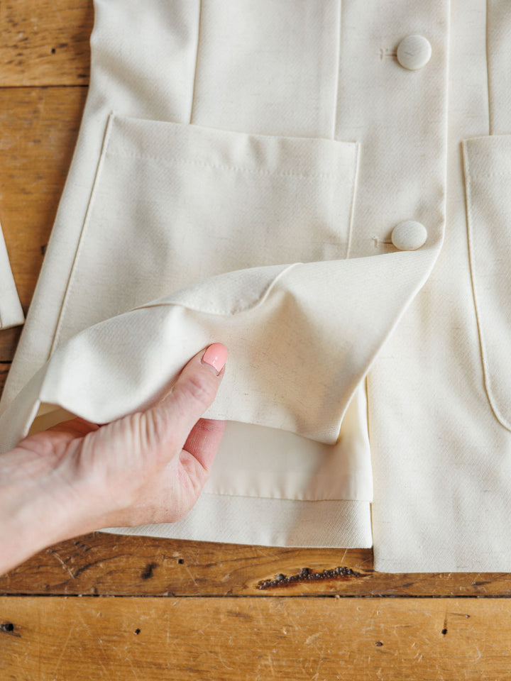 A hand  lifts the pocket of The Savona Set by Savannah Sevens western life&style, revealing inner construction details of the cream-colored sleeveless blazer lying flat on a wooden surface.