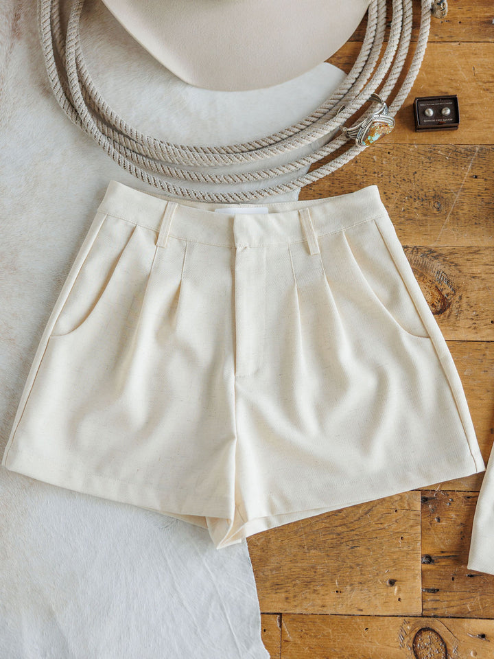 The Savona Set cream linen-blend shorts by Savannah Sevens western life{&}style, with front pleats and side pockets, are displayed flat on wood beside a beige hat, belt buckle, and white textured fabric.
