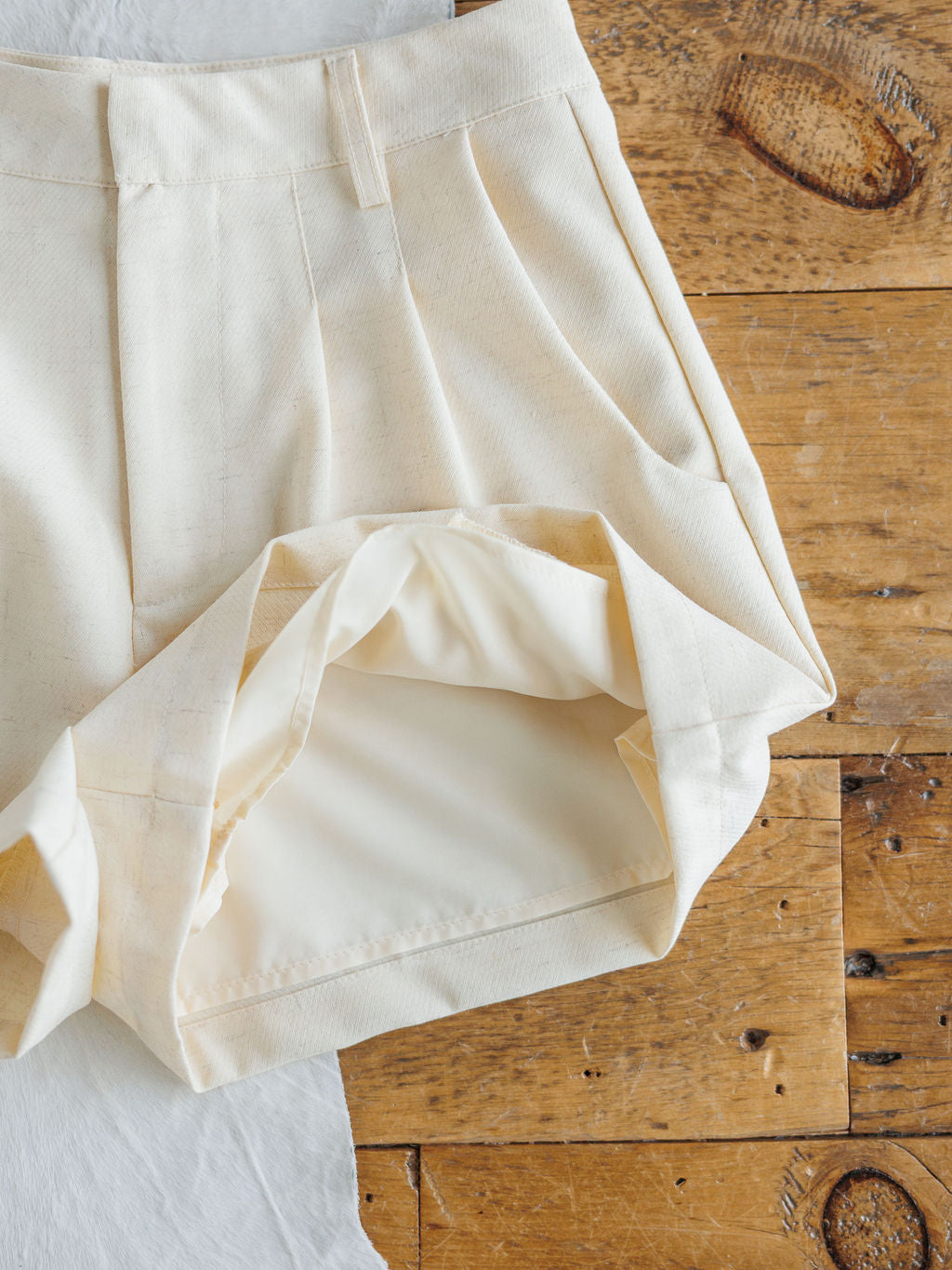 A close-up of The Savona Set cream-colored high waisted linen blend shorts by Savannah Sevens western life&style, with the hem flipped up to reveal the white inner lining, displayed on a rustic wooden surface.