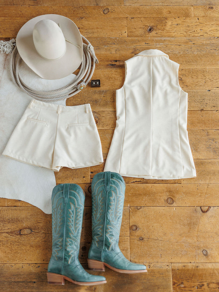 A Western-inspired look from Savannah Sevens western life{&}style features The Savona {Set}—a sleeveless blazer and high-waisted linen-blend shorts—styled with a cream cowboy hat and turquoise stitched cowboy boots.