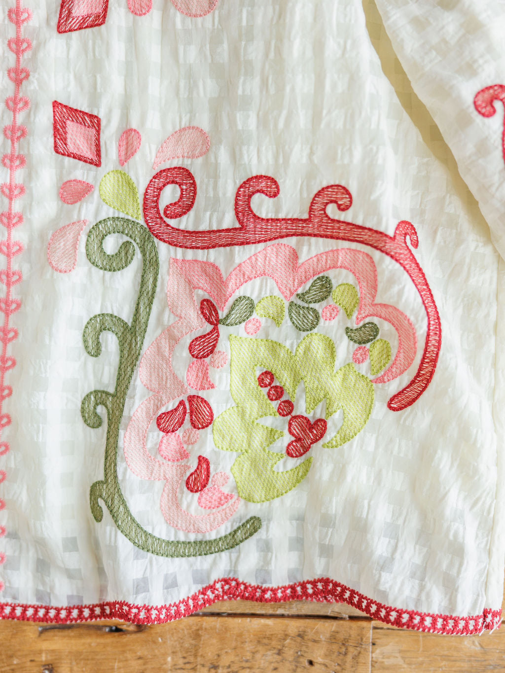 A close-up of The Elrosa {Set} from Savannah Sevens western life{&}style shows white fabric with vibrant floral and swirl embroidery in green, red, and pink, finished with red trim—suggesting a chic embroidered top & shorts set on wood.